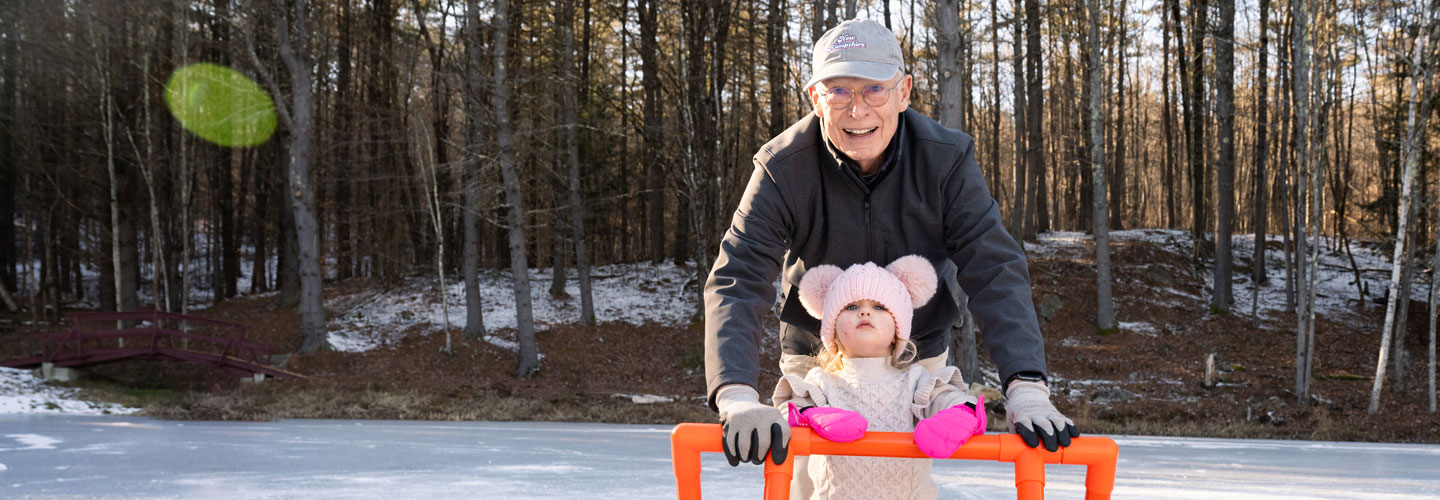 Photo of grandfather and young grand daughter leaning on an orange colored hockey goal with snowy background and trees